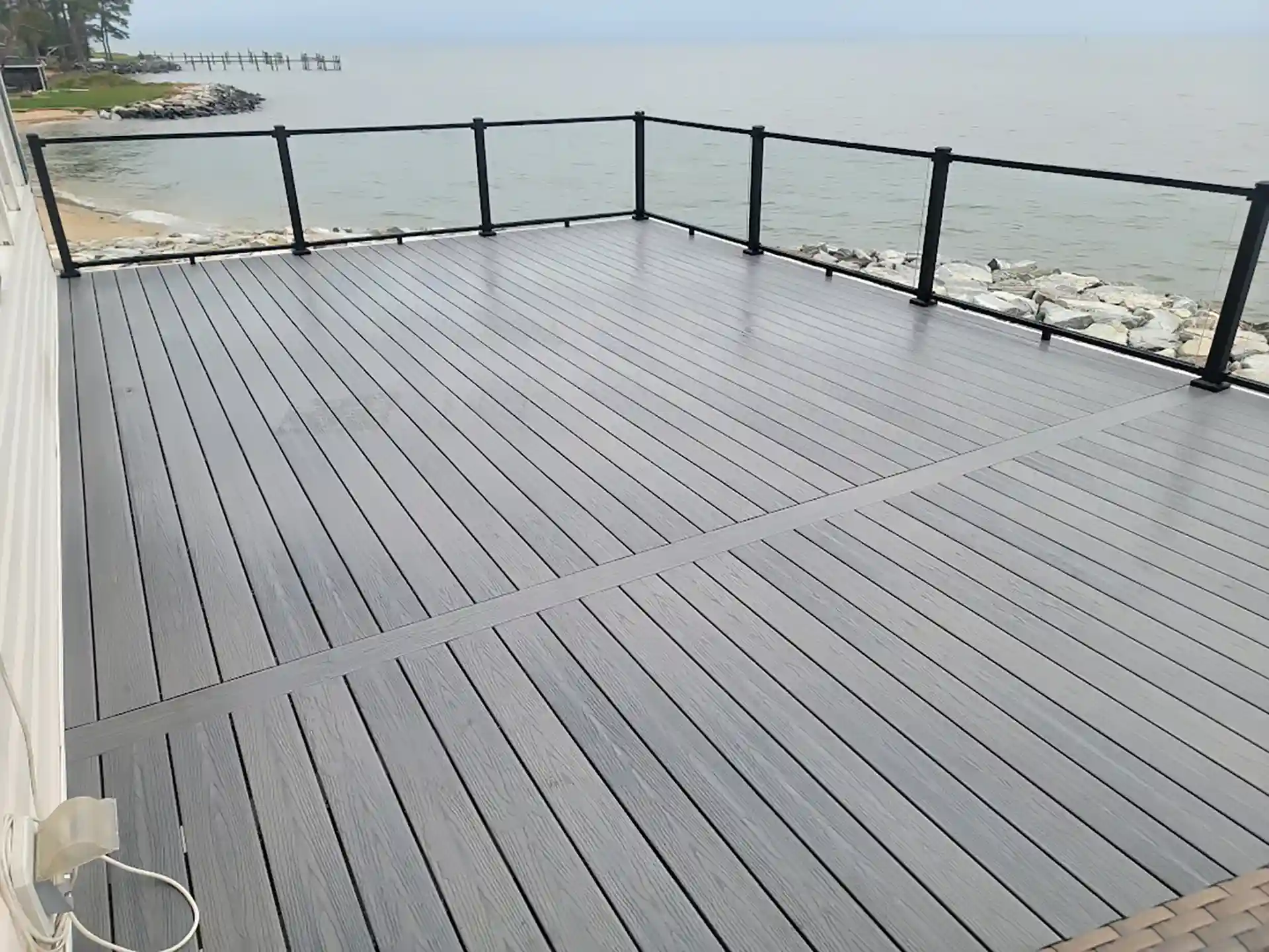 Gray composite waterfront deck with black metal and glass railing overlooking a rocky shoreline.
