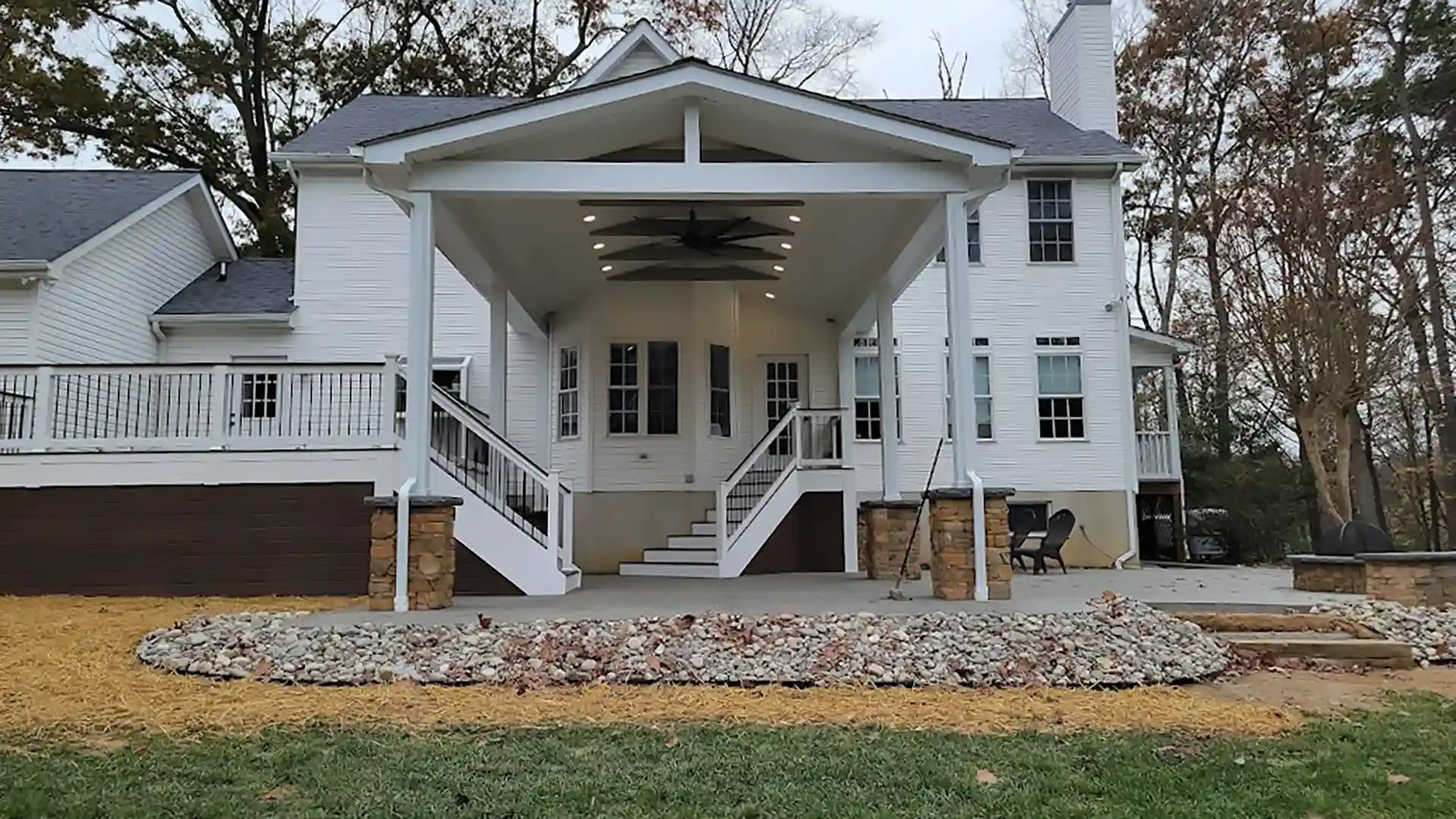 Covered back porch with gable roof, recessed lighting, large ceiling fan, and white columns leading to a patio outside a two-story house. 