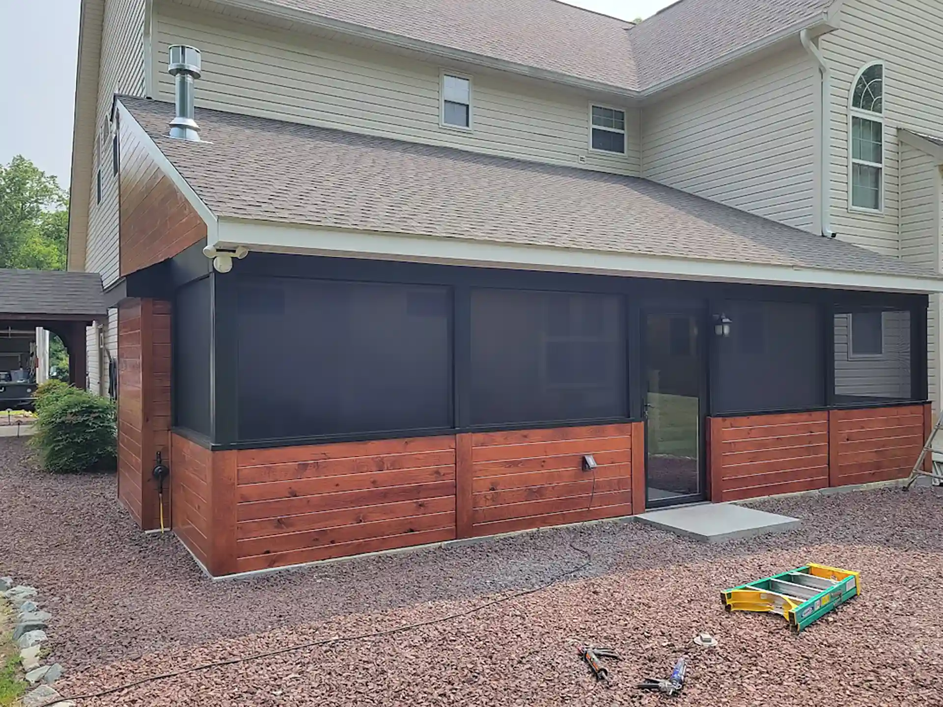 Screened-in porch with dark mesh panels, stained wood lower walls, and a sloped roof attached to the back of a beige two-story house. 