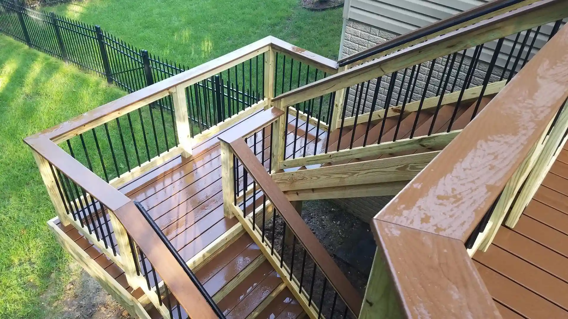 A wooden deck with stairs, black metal railings, and freshly applied sealant, leading to a grassy area. 