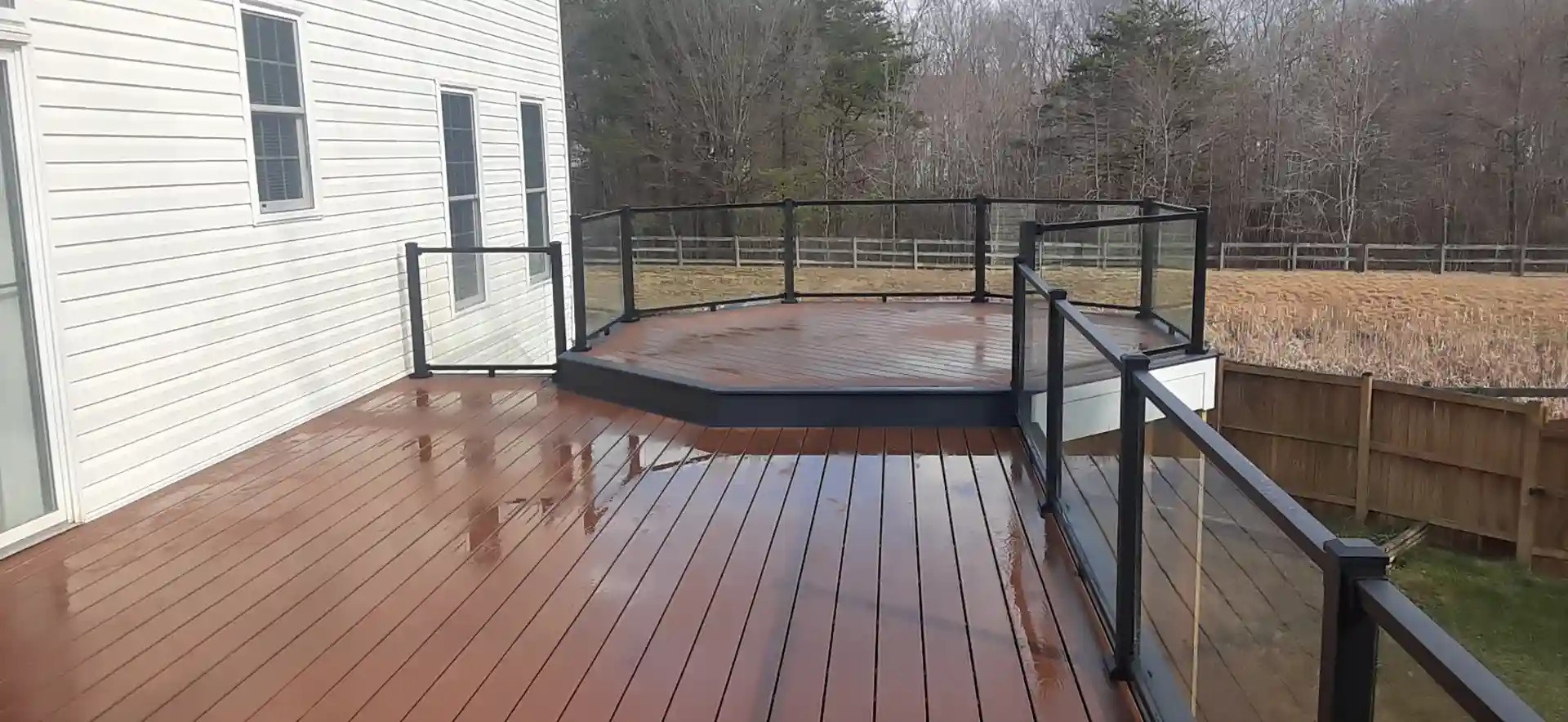A wooden deck with a glass railing, featuring an octagonal section, attached to a house with a fenced backyard. 