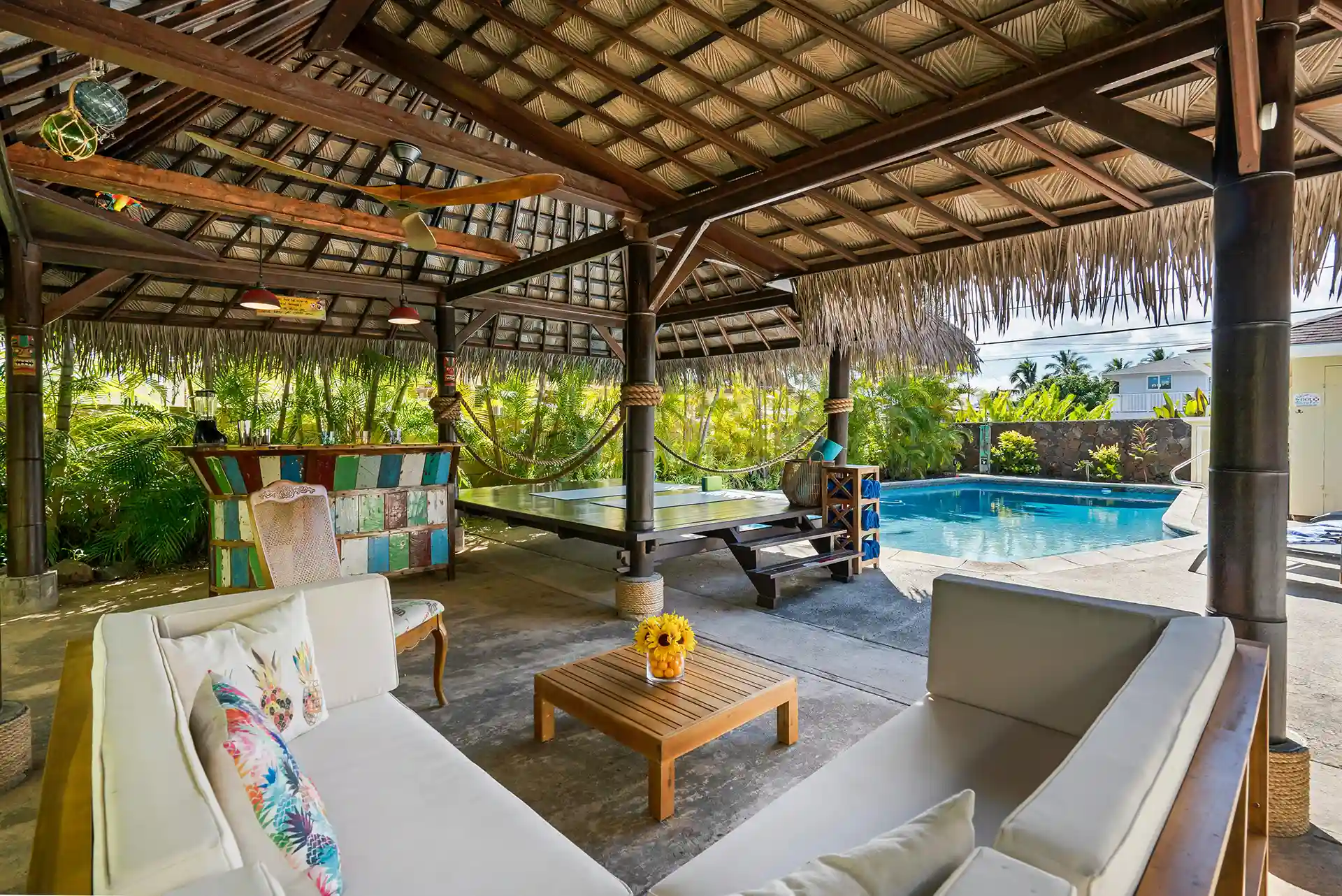 Outdoor tiki-style pavilion with wood framing, thatched roof, lounge seating, and a bar area next to a private swimming pool surrounded by tropical landscaping. 